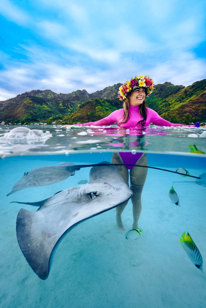 Person in a pink wetsuit with a flower crown interacting with stingrays underwater, with mountains in the background.
